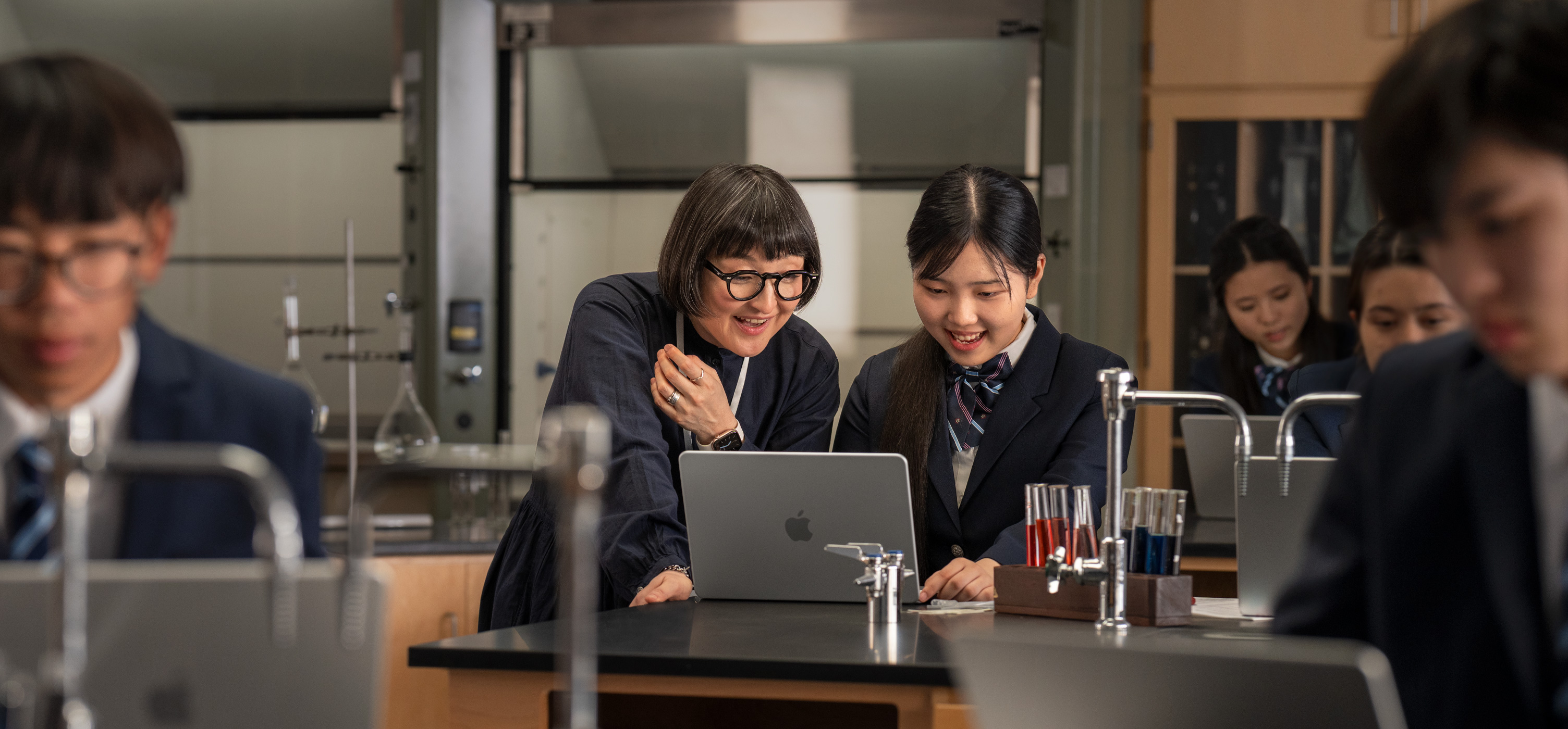 一位學(xué)生和一位教師在實驗室環(huán)境下互動，桌上放著打開的 MacBook，周圍滿佈試管和設(shè)備