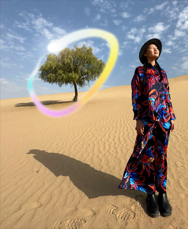 Bild einer Frau in einem mehrfarbigen Kleid, die in einer Wüstenlandschaft mit einem einsamen Baum im Hintergrund steht, der von einem leuchtenden Regenbogenverlauf umgeben ist, um die Apple Intelligence F?higkeit zum L?schen von Hintergrundobjekten zu zeigen