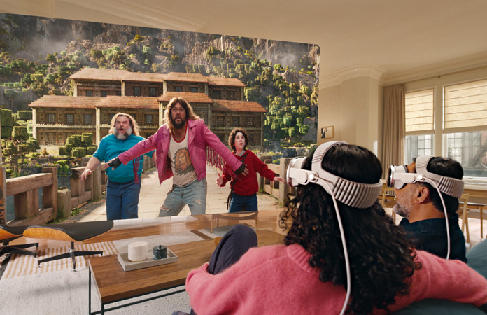 Zwei Personen, die beide eine Apple Vision Pro tragen, sitzen nebeneinander auf der Couch und schauen ?Ein Minecraft Film“, wobei sich der Bildschirm über den gesamten Raum erstreckt
